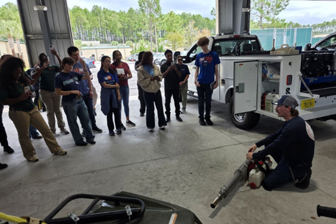 A group of students is watching a mosquito control professional demonstrating equipment.