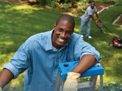 Young man mows lawn in background while man is on ladder clearing debris from house guttering in foreground.